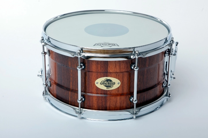 Snaredrum-13x7-Jarrah Nahaufnahme einer Snare-Drum aus Jarrah-Holz mit einem warmen, rötlich-braunen Finish. Die Trommelkessel sind mit verchromter Hardware ausgestattet, darunter Spannreifen, Lugs und Spannschrauben. Das Trommelfell zeigt eine mittige Verstärkung für zusätzliche Haltbarkeit
