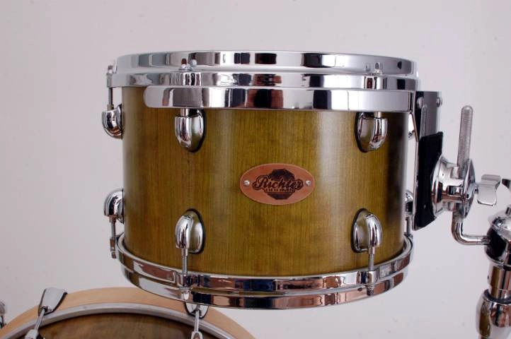Kirschholz Schlagzeug-gruen Nahaufnahme einer Tom-Tom-Drum mit grünlich-braunem Holzfinish und verchromter Hardware. Auf dem Kessel ist ein Metallbadge mit der Aufschrift 'Richter Drums' zu sehen. Die Trommel ist auf einem Stativ montiert, und ein weiteres Schlagzeugelement ist im unteren Bereich sichtbar.