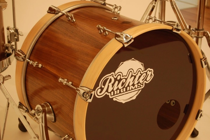 Nussbaum-Schlagzeug Nahaufnahme einer Bassdrum mit natürlichem Holzfinish und verchromter Hardware. Das Trommelfell trägt das Richter Drums-Logo in weißer Schrift auf dunklem Hintergrund. Teile weiterer Trommeln und Ständer sind im Hintergrund sichtbar.