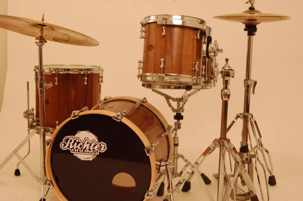 Schlagzeug-aus-Nussbaum. Schlagzeug-Set mit Bassdrum, Snare, Hängetom und Floor Tom, alle mit edlem Holzfinish und verchromter Hardware. Die Bassdrum trägt das Richter Drums-Logo auf dem Trommelfell. Zwei Becken sind auf stabilen Ständern montiert.