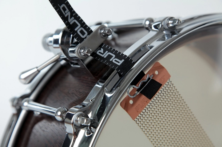 Snaredrum.wenge Nahaufnahme einer Snare-Drum mit Holzkessel, verchromter Hardware und sichtbaren Snare-Drähten unter dem Trommelfell. Ein schwarzer Gurt mit der Aufschrift 'PURE' ist am Snare-Mechanismus befestigt.